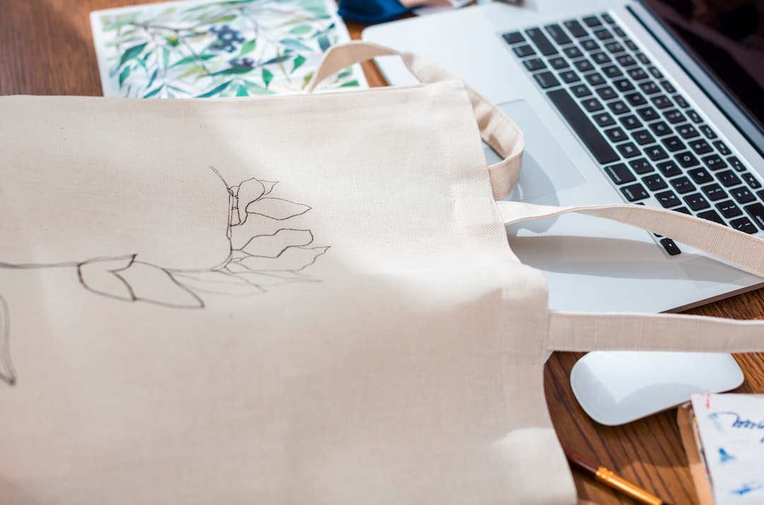 Kremowa płócienna torba z delikatnym szkicem liścia obok laptopa, myszki, ołówków i botanicznej grafiki na drewnianym biurku, co sugeruje kreatywność.
